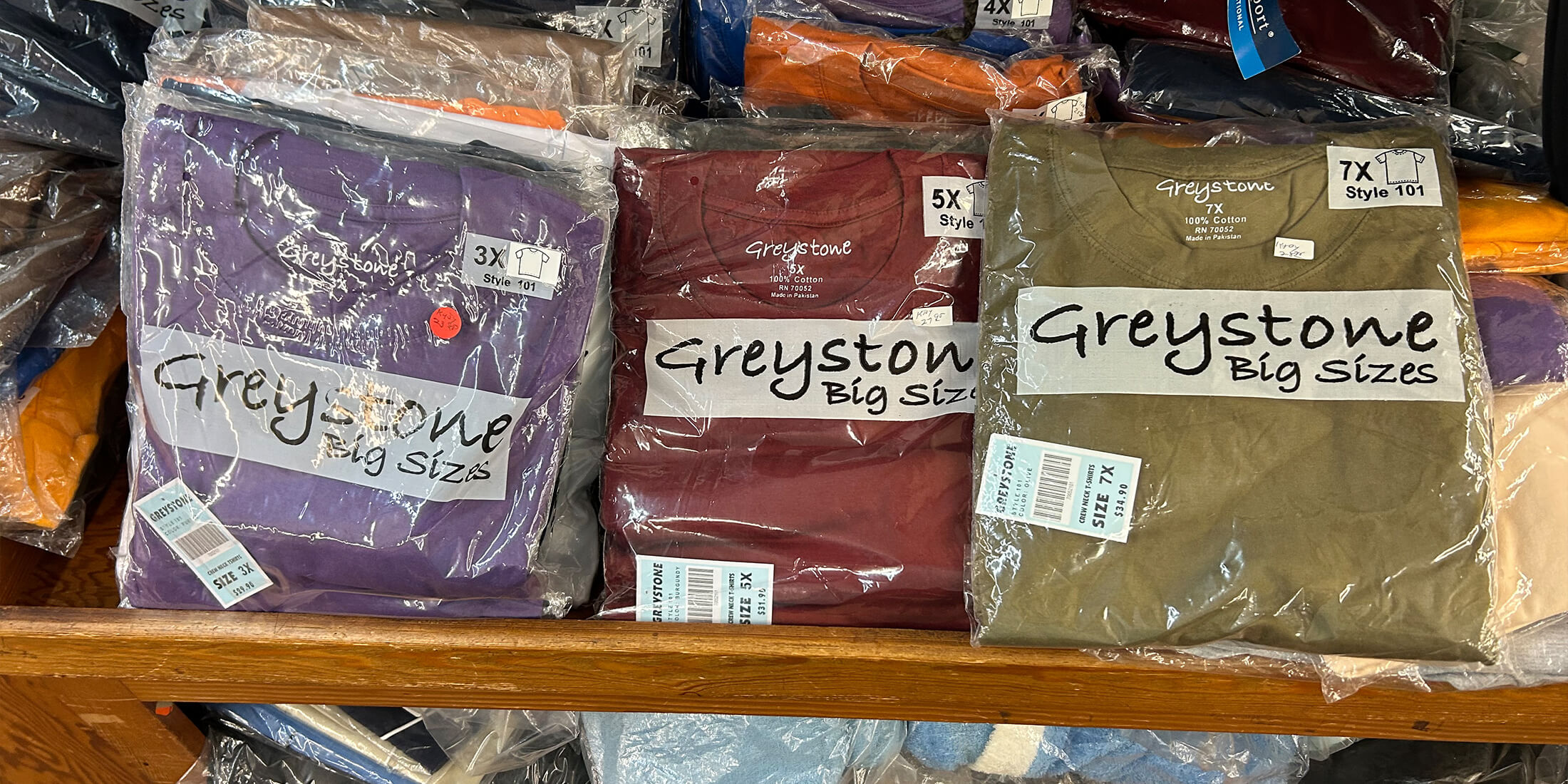 Stacks of folded Greystone Big Sizes crew-neck t-shirts in 3X, 5X and 7X on a shelf at Kornfeld's Department Store.